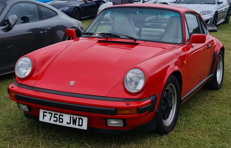 Guards Red 1988 Porsche 911 Carrera 3.2 coupé with impact bumpers and Fuchs wheels on grass at the Goodwood Festival of Speed, three-quarter front view