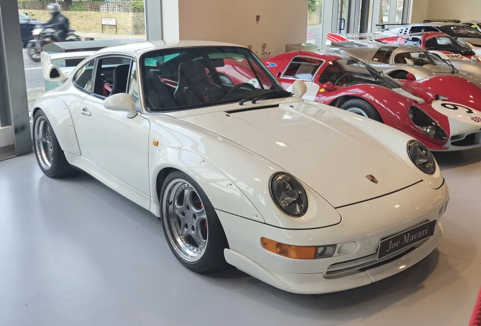 White Porsche 911 GT2 (993) with bolt-on arch extensions and biplane rear wing in a London showroom, three-quarter front view