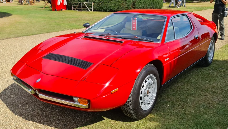 Red Maserati Merak on grass at a concours, three-quarter front view showing the open flying-buttress rear