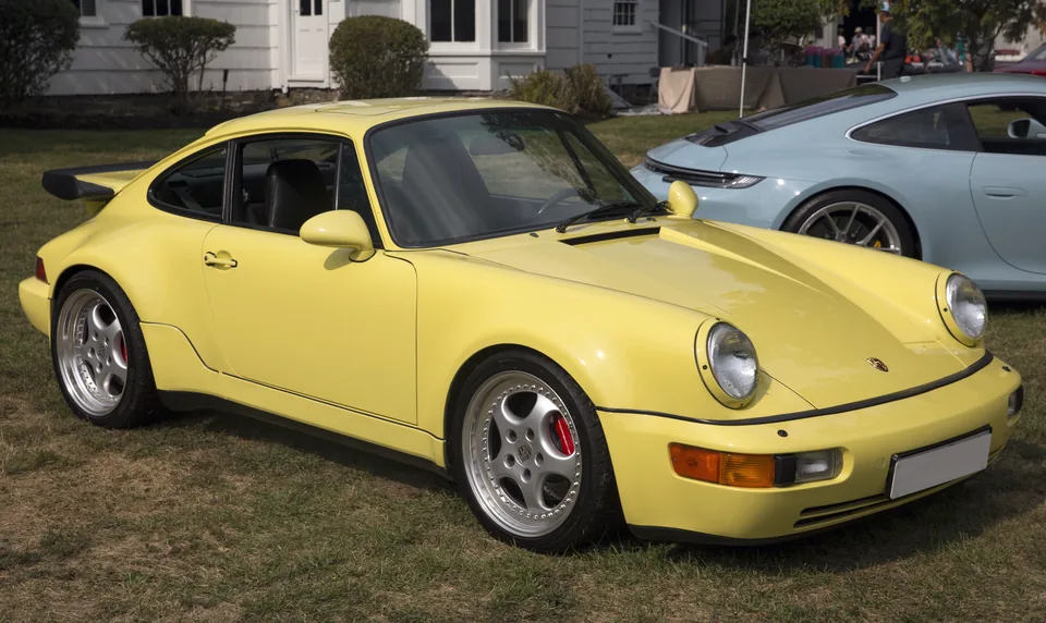 Summer Yellow 1991 Porsche 911 Turbo (964) with wide arches and tea-tray spoiler on grass at a Long Island cars-and-coffee, three-quarter front view